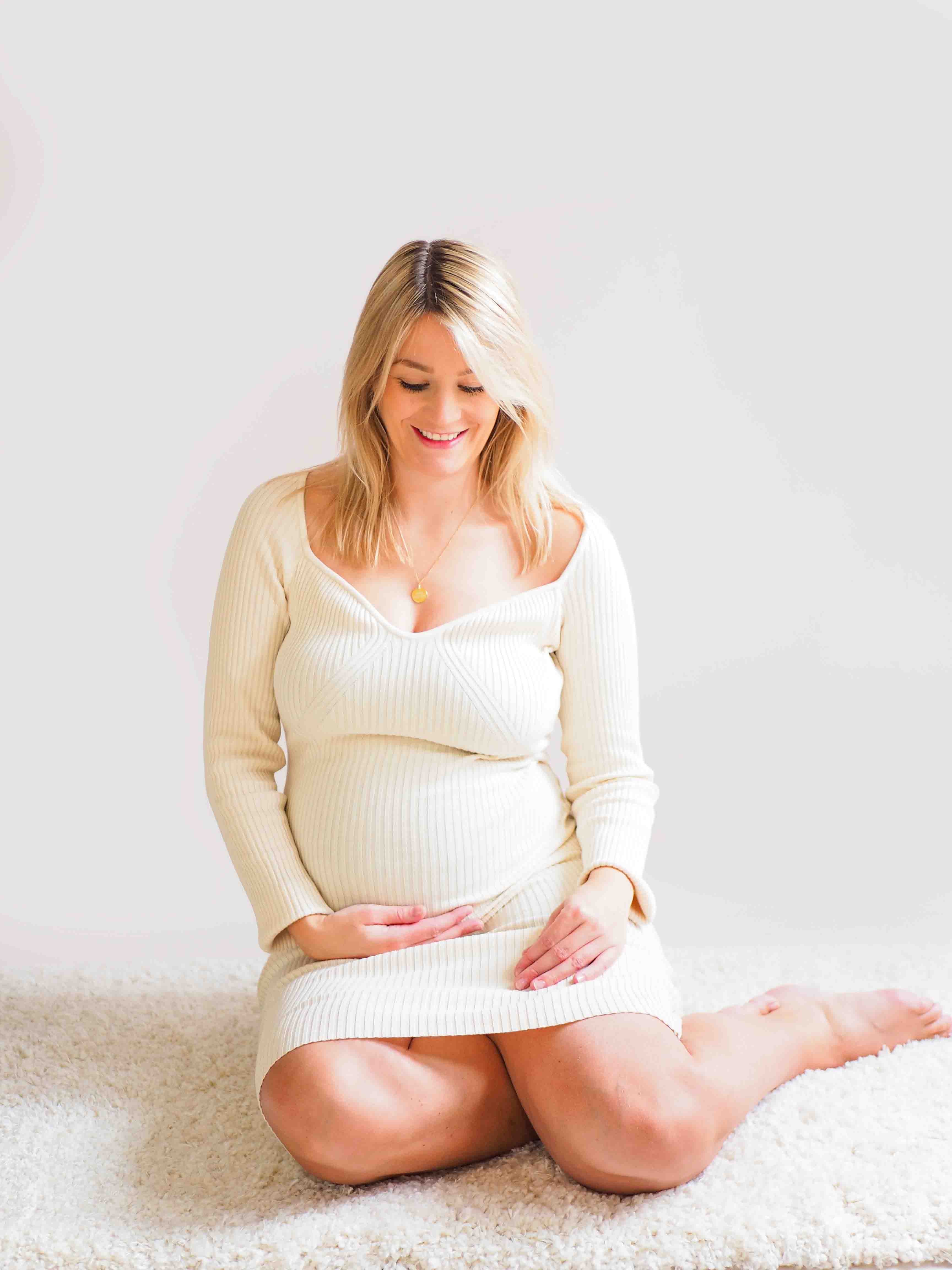 Een stralende zwangere vrouw staat in een prachtig omgeving omringd door zachte verlichting en een elegante achtergrond. Haar handen rusten liefdevol op haar ronde buik terwijl ze vol verwachting naar de camera glimlacht in de studio bij van Lente fotografie in Spijkenisse.