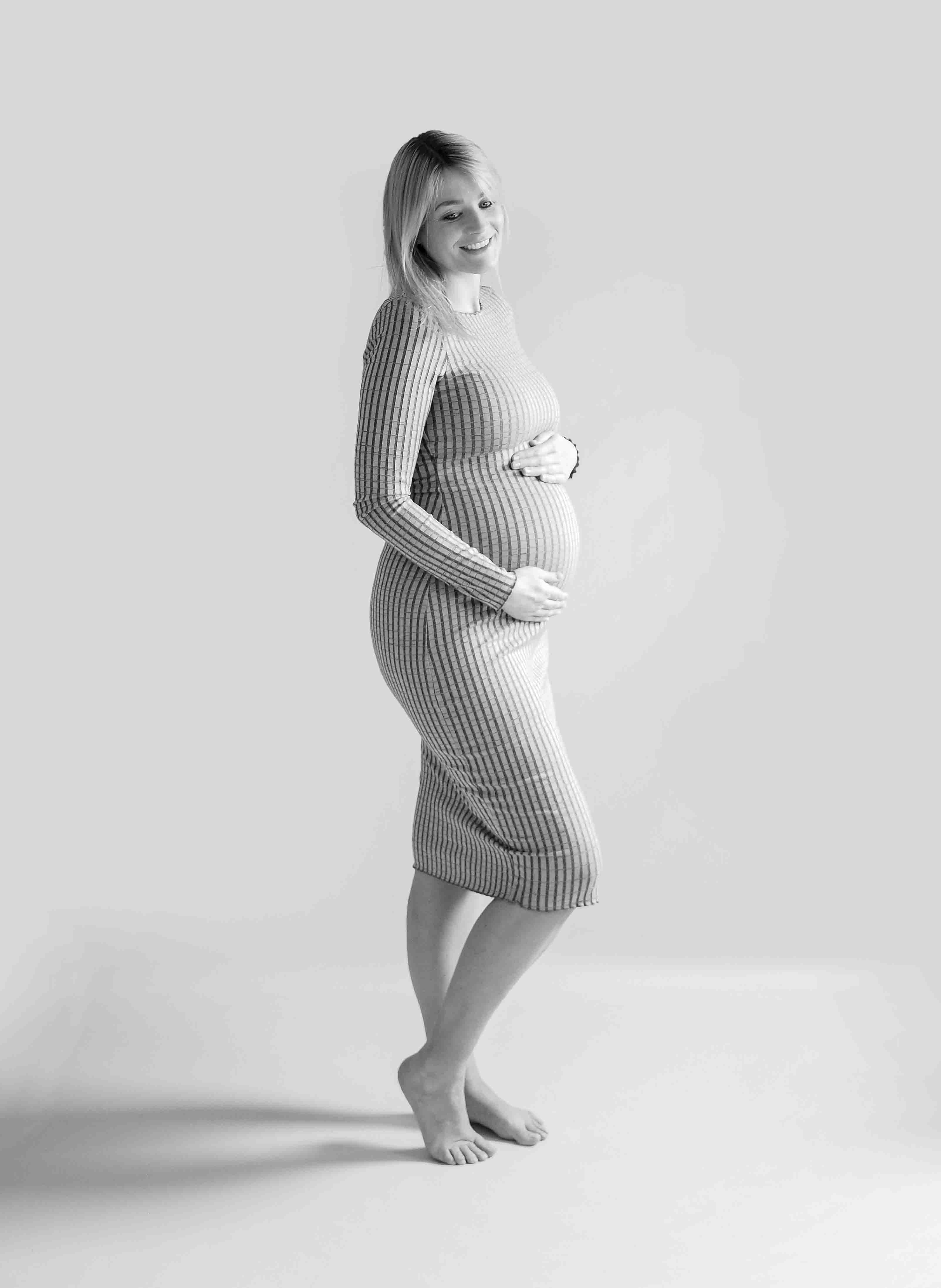 Een stralende zwangere vrouw staat in een prachtig omgeving omringd door zachte verlichting en een elegante achtergrond. Haar handen rusten liefdevol op haar ronde buik terwijl ze vol verwachting naar de camera glimlacht in de studio bij van Lente fotografie in Spijkenisse.