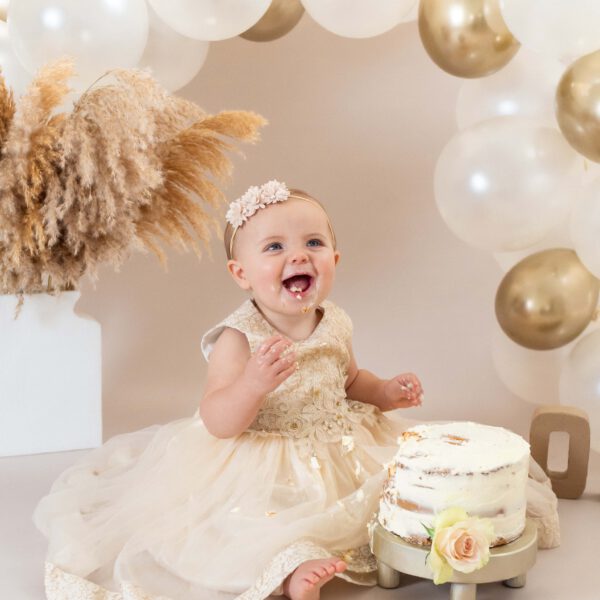 Een vrolijk baby meisje van 1 jaar straalt op de foto omgeven door taartkruimels, haar gezicht en handen bedekt met frosting, terwijl ze lachend en vol vreugde haar eerste verjaardagstaart verkent tijdens een cakesmash shoot in de studio bij van Lente fotografie in Spijkenisse.