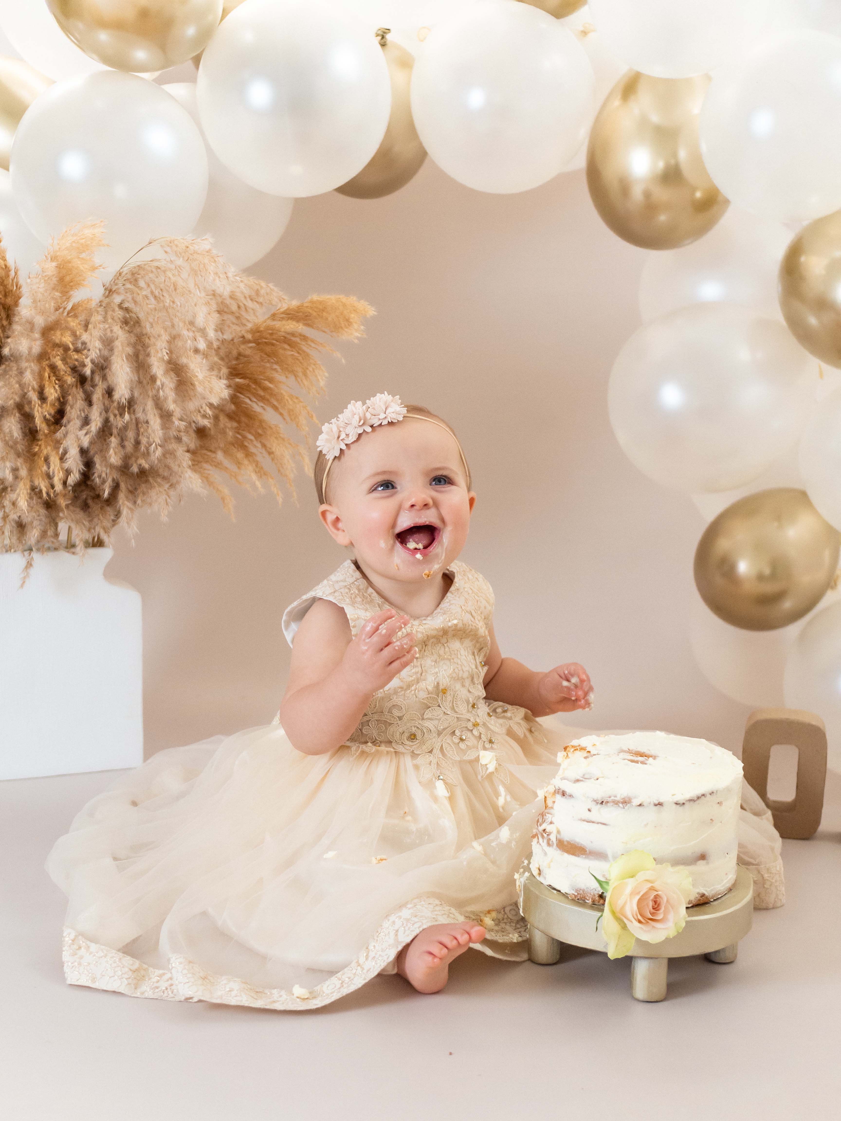 Een vrolijk baby meisje van 1 jaar straalt op de foto omgeven door taartkruimels, haar gezicht en handen bedekt met frosting, terwijl ze lachend en vol vreugde haar eerste verjaardagstaart verkent tijdens een cakesmash shoot in de studio bij van Lente fotografie in Spijkenisse.