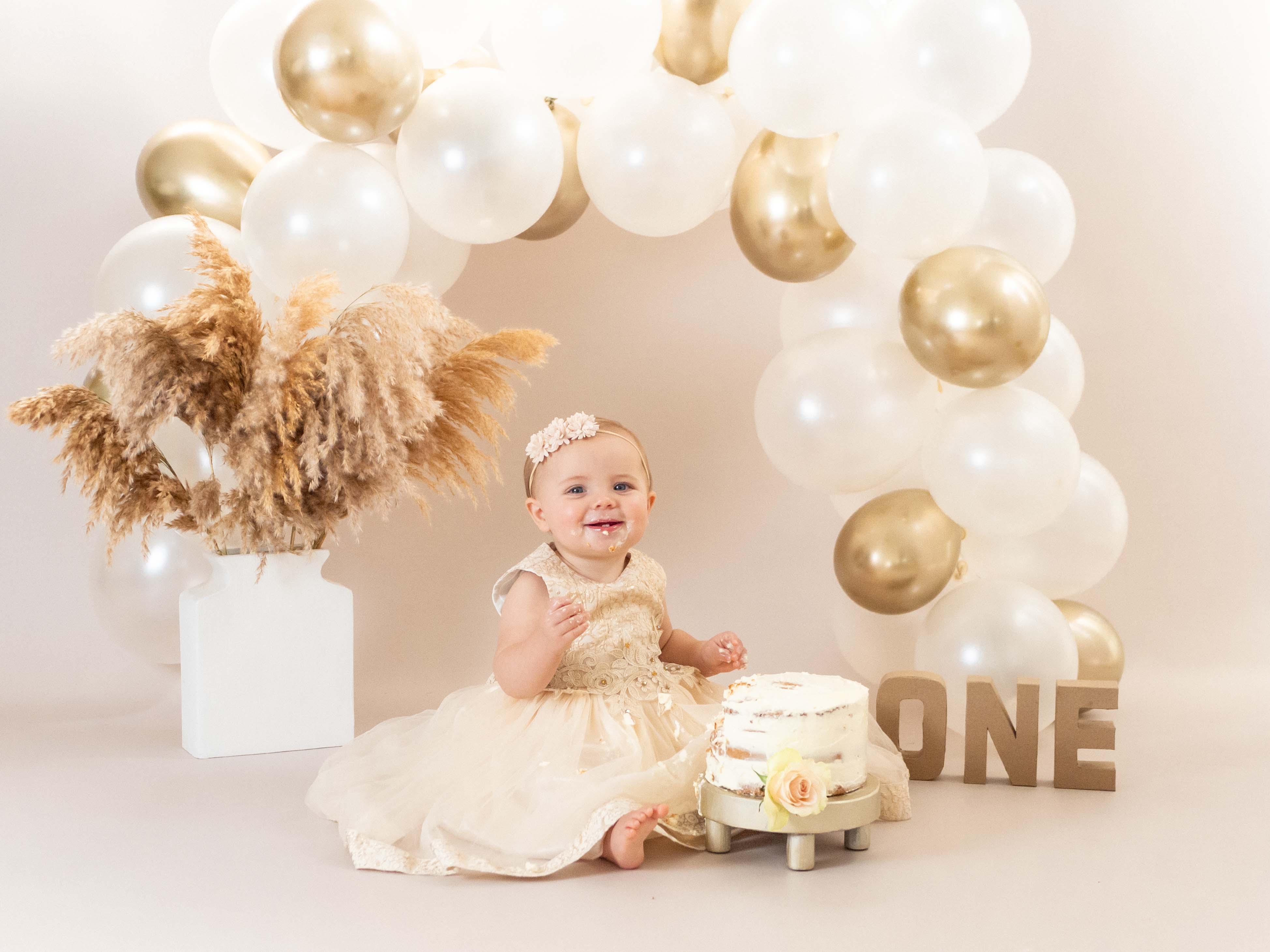 Een vrolijk baby meisje van 1 jaar straalt op de foto omgeven door taartkruimels, haar gezicht en handen bedekt met frosting, terwijl ze lachend en vol vreugde haar eerste verjaardagstaart verkent tijdens een cakesmash shoot in de studio bij van Lente fotografie in Spijkenisse.