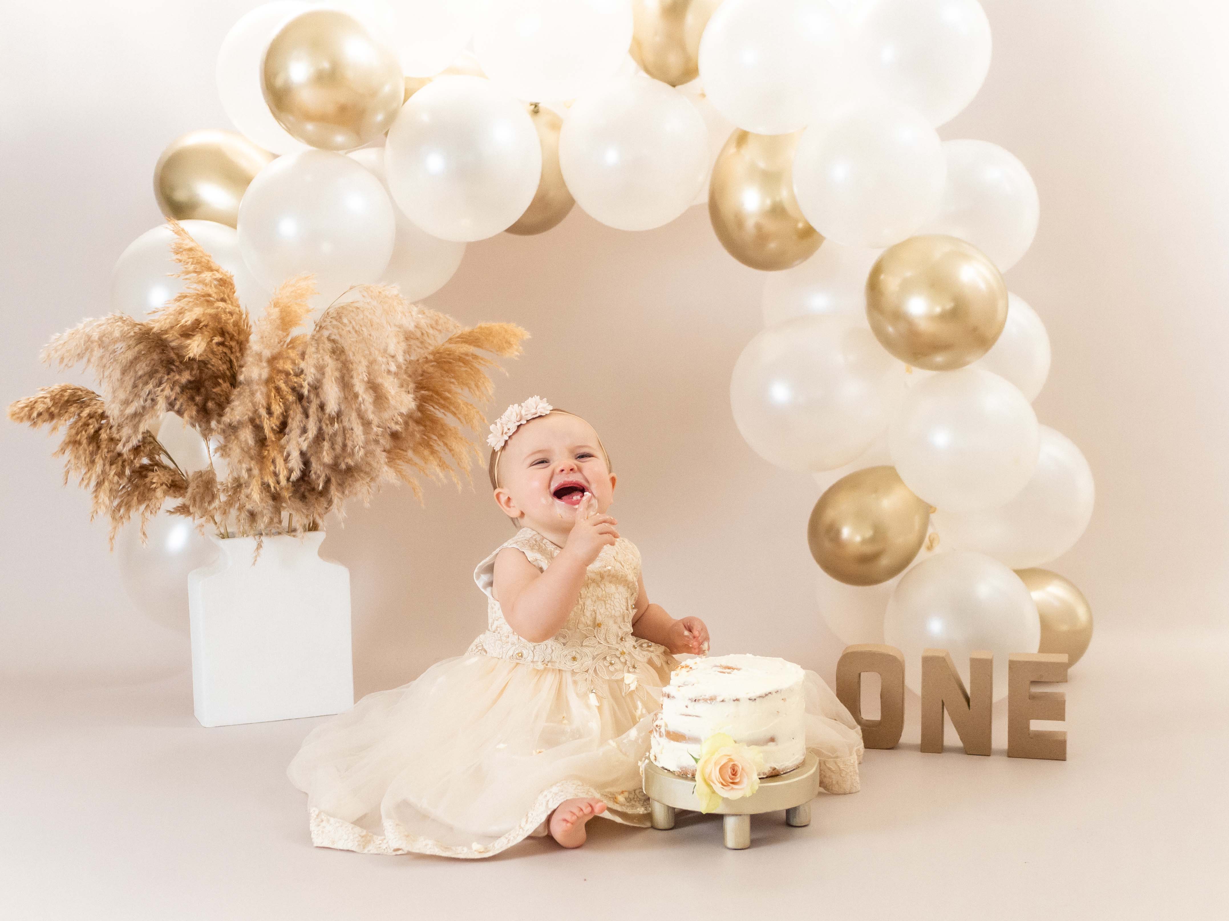 Een vrolijk baby meisje van 1 jaar straalt op de foto omgeven door taartkruimels, haar gezicht en handen bedekt met frosting, terwijl ze lachend en vol vreugde haar eerste verjaardagstaart verkent tijdens een cakesmash shoot in de studio bij van Lente fotografie in Spijkenisse.