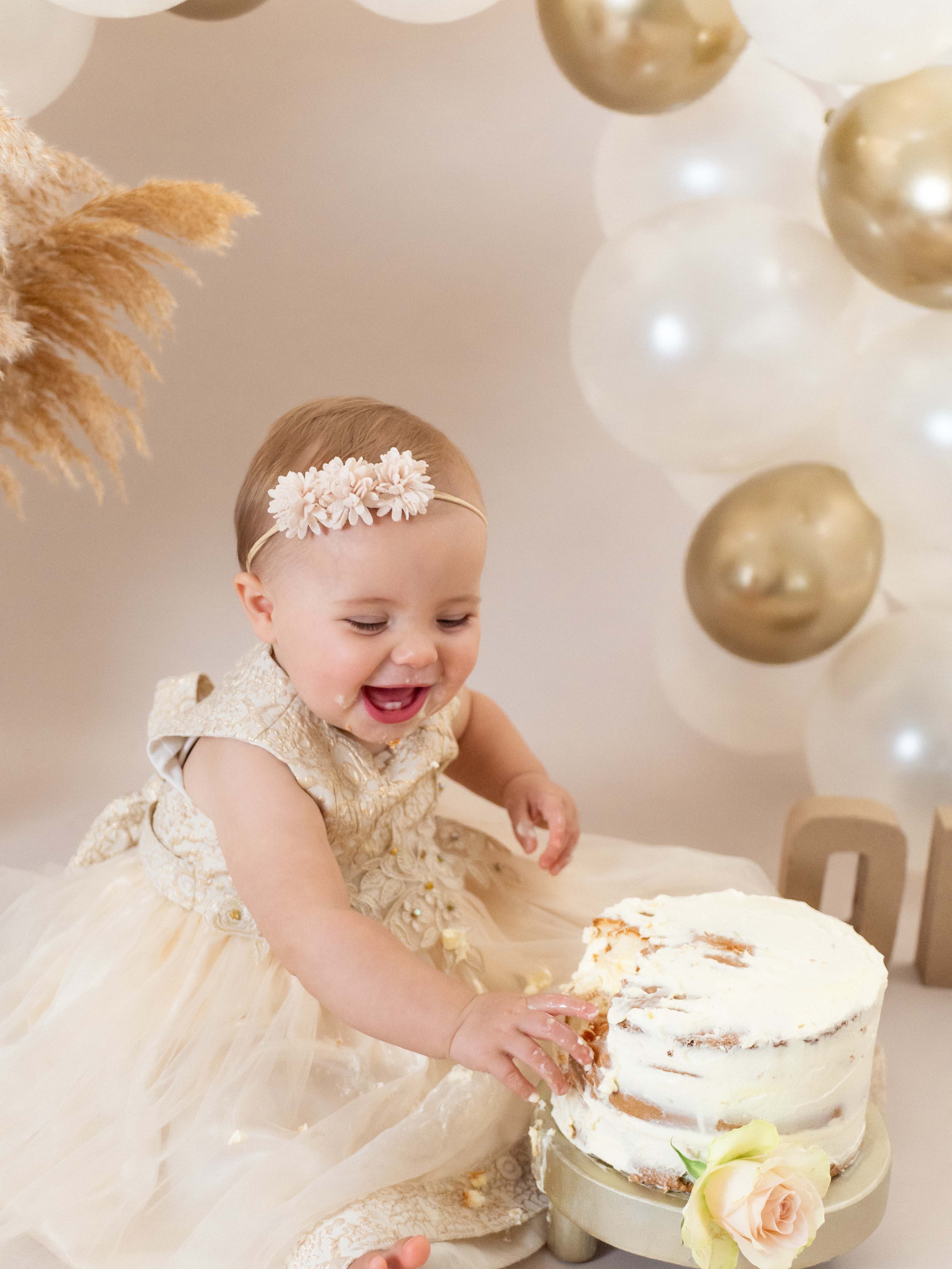 Een vrolijk baby meisje van 1 jaar straalt op de foto omgeven door taartkruimels, haar gezicht en handen bedekt met frosting, terwijl ze lachend en vol vreugde haar eerste verjaardagstaart verkent tijdens een cakesmash shoot in de studio bij van Lente fotografie in Spijkenisse.